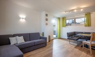 A bright living room with a gray sofa and a dining table. The windows are decorated with green curtains, and the floor is made of wood.