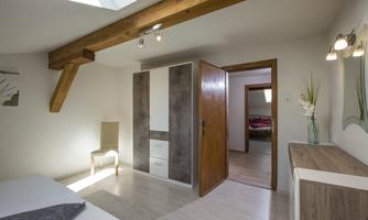A bright room with a window and a wooden beam on the ceiling. There is a wardrobe, a chair, and a dresser.