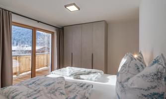 A modern bedroom with a large window and balcony. The walls are light, and the bedding is in soft colors.