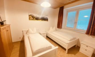 A simple bedroom with two single beds and a cozy wooden shelf. The room has a window with a view of the mountains and warm lighting.