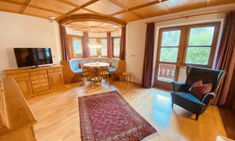 A cozy living room with wooden paneling and a large window. There is a dining table, chairs, and a television.