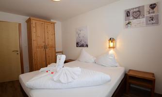 A cozy bedroom with a double bed and a wooden wardrobe. The walls are adorned with decorations and a picture, and a lamp provides pleasant light.