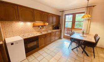 A modern kitchen with wooden cabinets and a dining area. Large windows provide plenty of daylight.