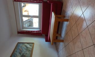 A simple living room with a red sofa and a wooden table. Two windows provide daylight, and a painting hangs on the wall.