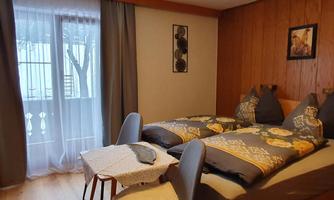 A cozy bedroom with two beds, a small table, and a view of the balcony. The walls are made of wood and there are curtains that let in light.