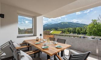 An elegant balcony with a large wooden table and chairs, beautifully set. In the background, green mountains and a clear sky can be seen.