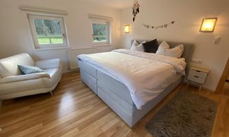 A cozy bedroom with a large bed and hardwood floor. Next to the bed is a comfortable armchair and windows with a view to the outside.