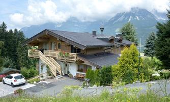 A charming wooden house amidst green meadows and mountains. The surroundings are picturesque with a blue sky and clouds.
