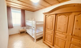 A simple room with a bunk bed and a wooden wardrobe. Bright walls and a window provide natural light.