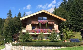 A charming wooden house with balconies full of flowers and a sunny sky. Surrounded by trees and a well-maintained green area.