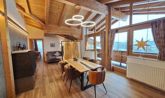 A modern living room with large windows and a dining area. Wooden ceilings and floors give the space a cozy atmosphere.
