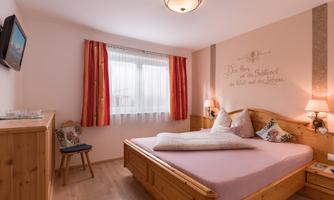 A cozy bedroom with a large wooden bed and windows with curtains. A small television is mounted on the wall, and there are decorative pillows on the bed.