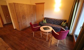 A cozy living area with a sofa and two red chairs. The room features a warm wooden floor and soft lighting.