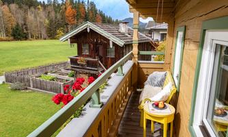 A cozy balcony view with a yellow table and chairs. In the background, there is a traditional wooden house and a green meadow.