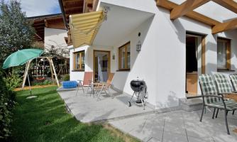 A cozy terrace with garden furniture and a barbecue. In the background, there is a playset for children and the sun is shining slightly.