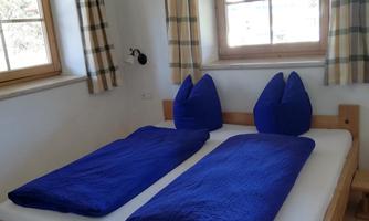 A cozy bedroom with two single beds and blue bedspreads. The room has large windows with checked curtains and plenty of daylight.