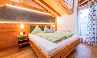 A cozy bedroom with wooden decor and a large bed. The walls are light, and there is plenty of daylight through the window.
