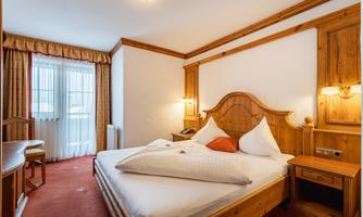 A cozy hotel room with wooden-paneled walls and a double bed. The room is bright and furnished with a table and chairs.