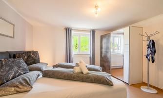 A modern bedroom with two beds, cozy pillows, and a window front. The room is bright and inviting, with a coat rack in the corner.