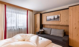 A cozy bedroom with a large bed and a gray sofa. The walls are made of wood and there are large windows with curtains.