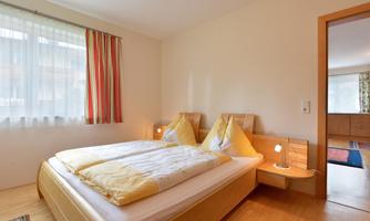 A bright bedroom with a double bed and two pillows. The room has large windows and simple decoration.