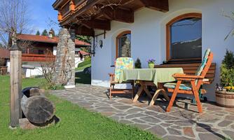 A cozy outdoor area with a table and chairs. In the background, green meadows and a traditional house can be seen.
