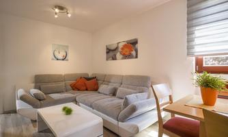 A cozy living room setup with a grey couch and a white table. Windows with blinds and decorative pictures on the wall give the room an inviting atmosphere.