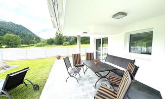 A cozy outdoor area with a table and several chairs. In the background, green meadows and mountains can be seen.