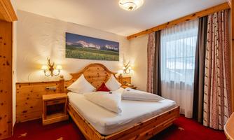 A cozy bedroom with a wooden bed and a red carpet area. Pictures hang on the walls and the windows let in plenty of daylight.