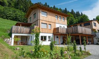 A modern wooden house with a large balcony and a well-maintained garden. In the background, green hills and trees are visible.