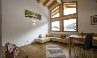A bright and modern living corner with large windows and plenty of natural light. There is a cozy seating area and a dining table.
