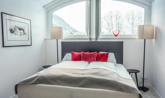 A cozy bedroom with a large bed and red pillows.  
Two lamps stand beside the bed and a window offers a view of nature.