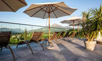 A relaxing area with lounge chairs and sun umbrellas on a terrace. In the background, mountains and a clear sky can be seen.