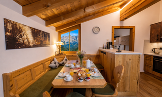 A welcoming dining area with wooden furniture and a large tabletop. On the table, breakfast dishes and drinks are arranged, while a beautiful mountain landscape can be seen through the window.