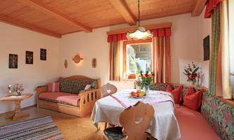 A cozy living space with a wooden ceiling and colorful furniture. A round table is decorated with flowers, and large windows let in plenty of light.