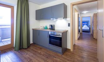 A modern kitchen with gray cabinets and wooden floor. In the background, a bright room with a bed can be seen.