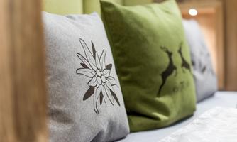Two decorative cushions on a bed, one gray with a floral pattern and the other green with a deer motif. In the background, a modern environment with wooden details is visible.