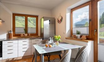 A modern kitchen with wooden windows and a table for four people. In the background, green mountains and a clear sky are visible.