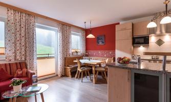 A cozy kitchen with a dining area and a living room. The walls are decorated in warm colors, and large windows offer a view of the greenery.