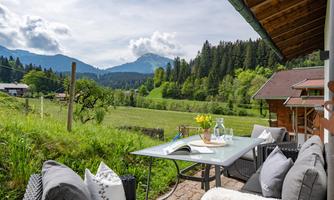A cozy terrace with a table and chairs, surrounded by an idyllic landscape. In the background, there are green hills and forests, while the mountains touch the sky.