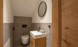A modern bathroom with a white toilet and a wooden washbasin. The walls are designed with gray tiles and wooden details.