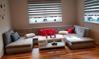 A cozy living room with an L-shaped couch and red pillows. The walls are light and the windows have blinds that let in light.