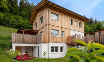 A modern wooden house in a green environment. The garden is equipped with loungers and surrounded by trees.