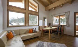 A modern living room with wooden ceilings and large windows. There is a cozy sofa, a dining area, and a rug on the floor.