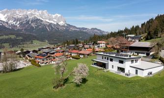 A picturesque mountain landscape with snow-capped peaks in the background. In the foreground, there are modern houses and a green meadow.