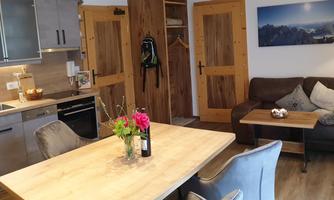 A cozy living kitchen with a wooden table, chairs, and a modern kitchen. In the room, there is a sofa and a decorative plant on the table.
