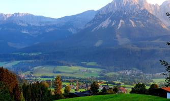 A picturesque mountain landscape with tall mountains and a clear sky. Below stretches a green valley with scattered houses and forests.