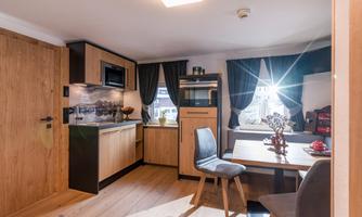 A modern, well-furnished kitchen room with wooden furniture and a dining table. Sunlight streams through the windows, creating an inviting atmosphere.