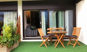 A cozy balcony with a wooden table and chairs. In the background, there is a window front and a green lawn area.