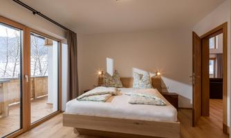 A modern bedroom with a large bed and two pillows. Large windows offer a view of the terrace and let in a lot of light.